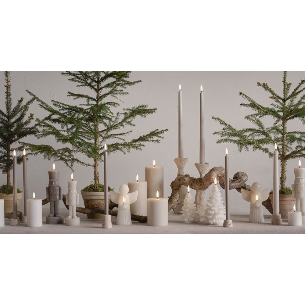 A collection of minimalist winter and Christmas candle holders and candles, including white pillar candles, angel figurines, driftwood horse holders, and small white tree ornaments, arranged on a shelf with potted pine trees in the background.
