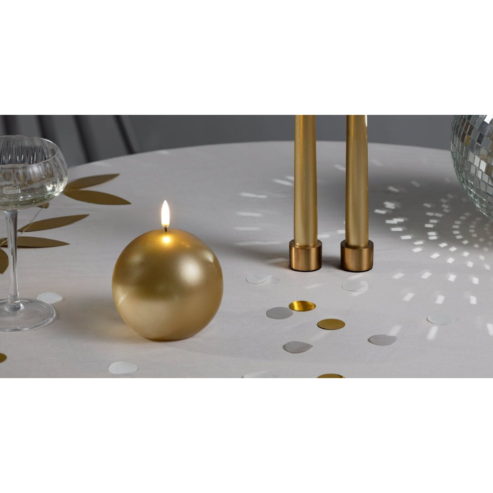 A gold spherical candle with a lit flame sits on a white tablecloth decorated with gold leaf motifs, alongside two gold taper candle holders and a glass goblet.