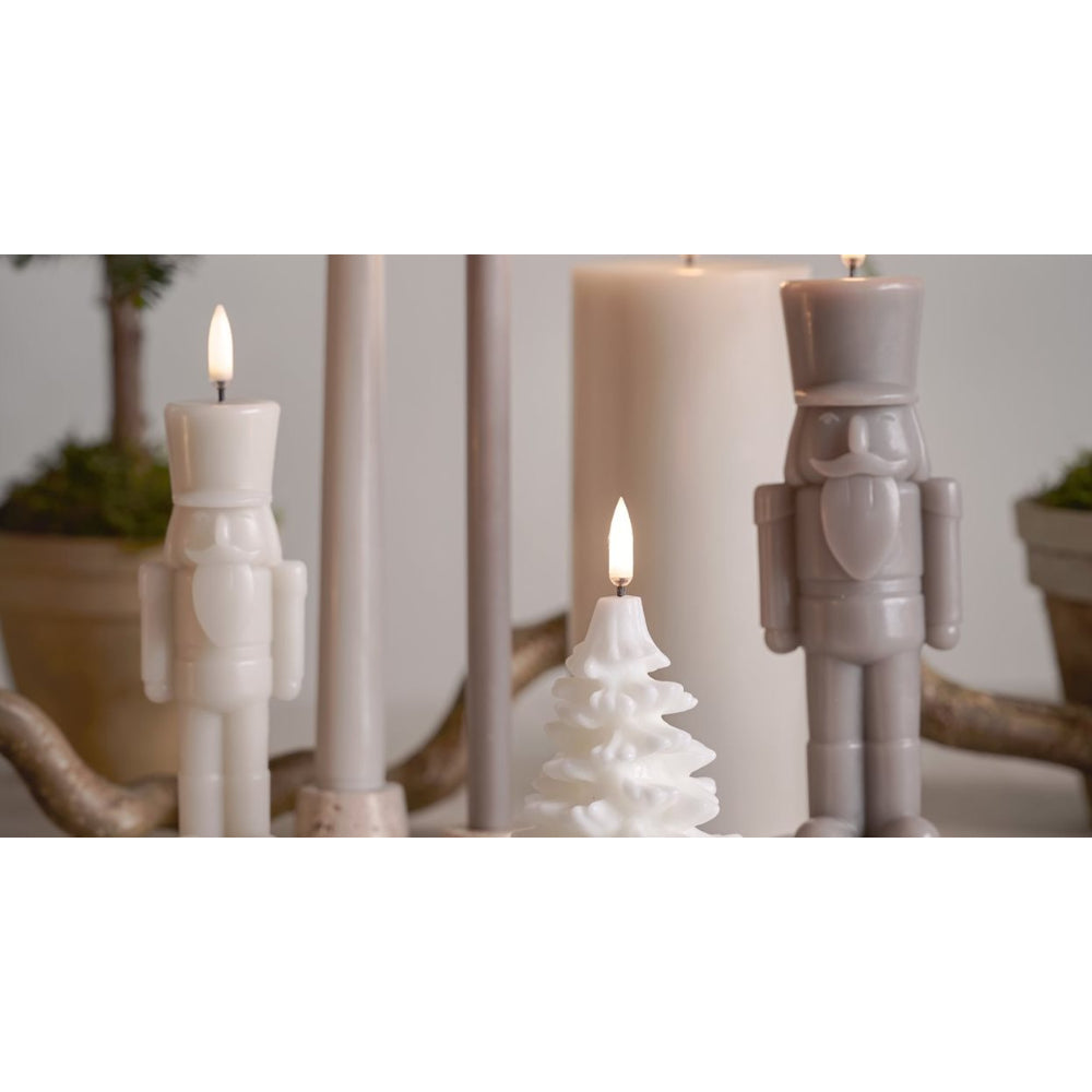 A set of decorative holiday candles, including two nutcracker-shaped candles in off-white and gray, a small white Christmas tree candle, and a large white pillar candle, styled on a rustic branch holder.