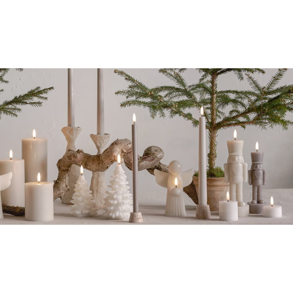 A styled holiday tabletop arrangement with multiple white and gray pillar candles, small white Christmas tree ornaments, a rustic driftwood centerpiece, and a potted evergreen tree against a light neutral wall.