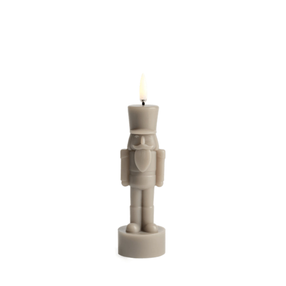 A nutcracker-shaped candle in a light taupe-beige color, shown lit with a small flame against a solid white background.