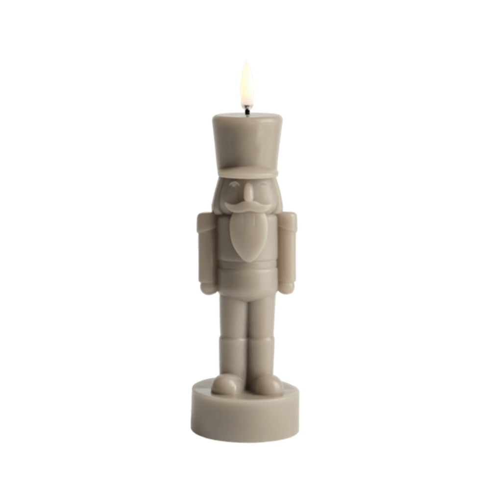 A nutcracker-shaped candle in a warm taupe-beige color, shown lit with a small flame against a solid white background.