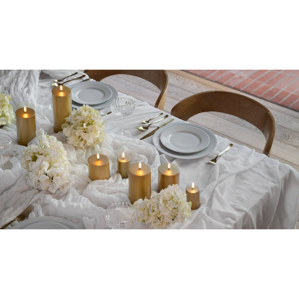 A styled dining table setting with white tablecloth, white plates, gold pillar candles, and white floral arrangements, with wooden chairs partially visible.