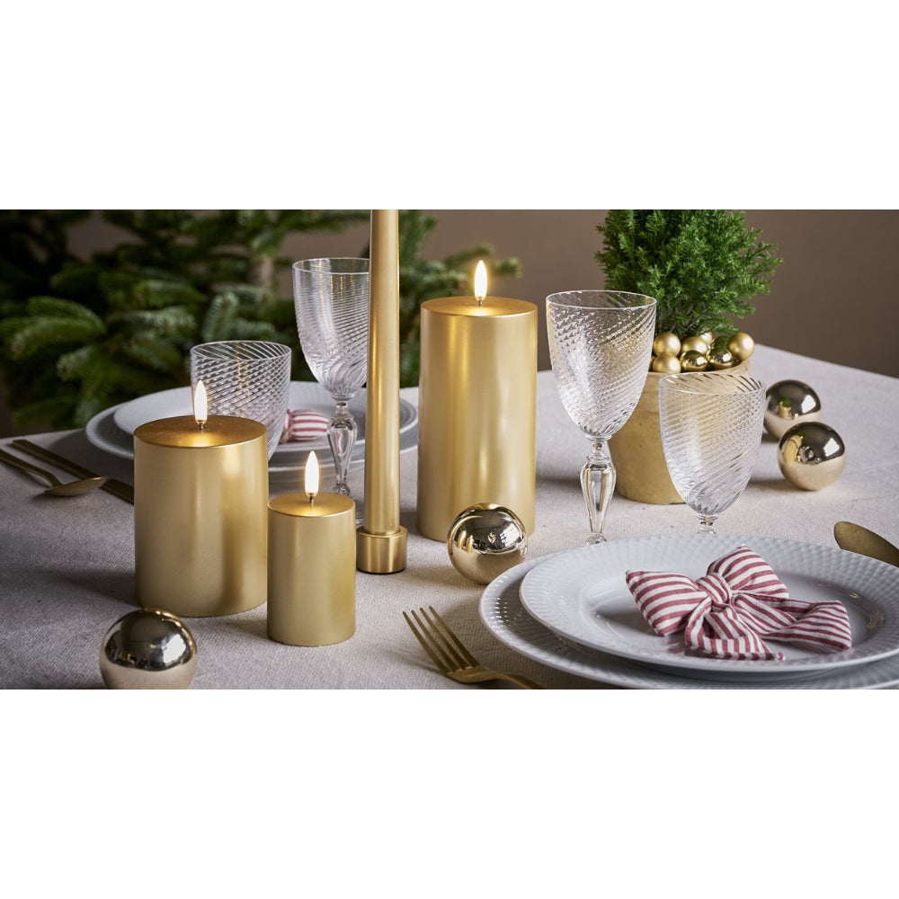 A styled holiday table setting featuring gold pillar candles, clear textured wine glasses, white plates, gold decorative balls, and a red-and-white striped napkin, arranged on a light gray tablecloth.