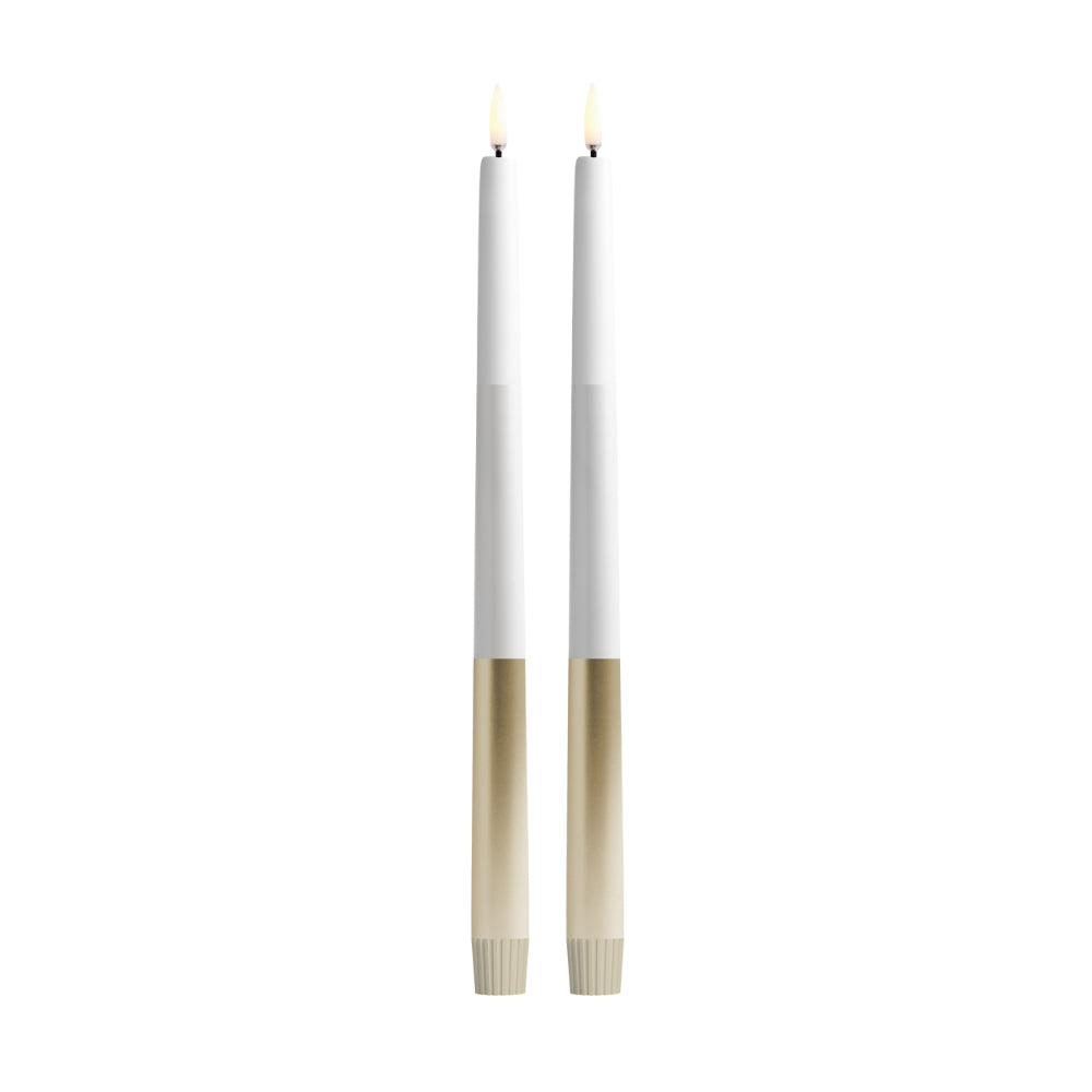 Two slim white taper candles with metallic gold lower sections and small lit flame tips, shown upright on a plain white background.