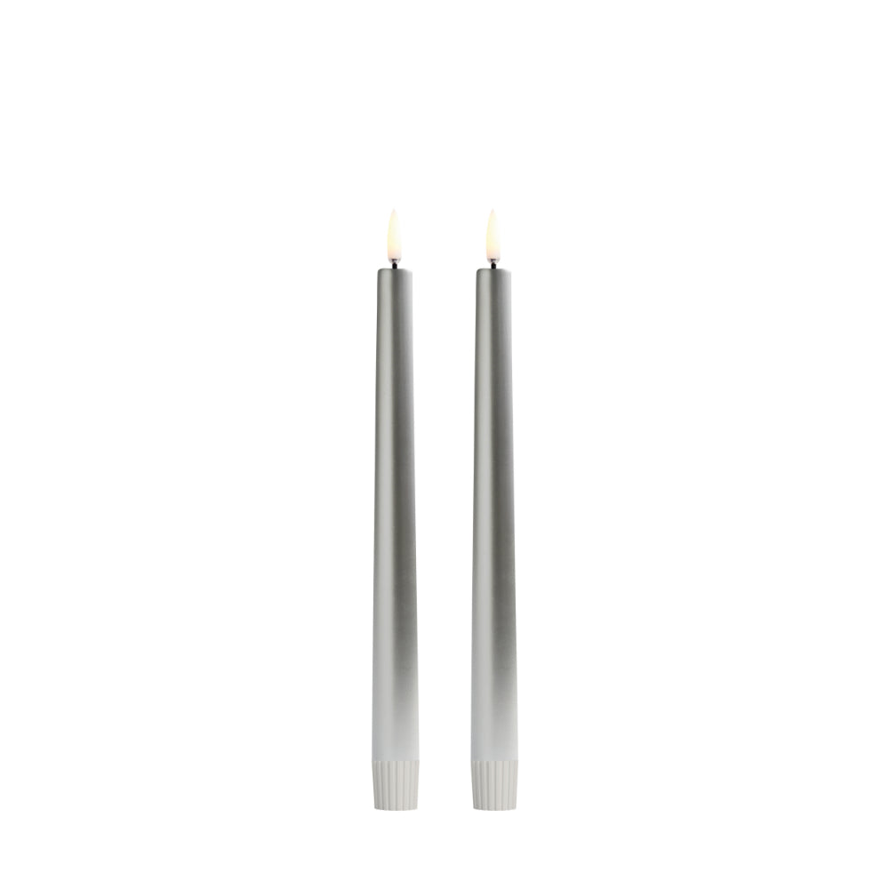Two slim silver taper candles with lit flame tips, standing upright side by side on a plain white background.