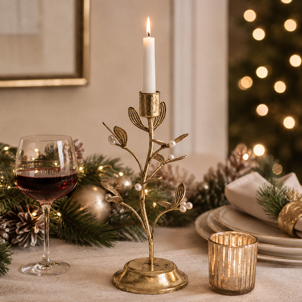 A gold holiday tree branch candle holder with a lit white taper candle, styled on a table with pine branches, a glass of red wine, plates, and a warm blurred Christmas tree in the background.