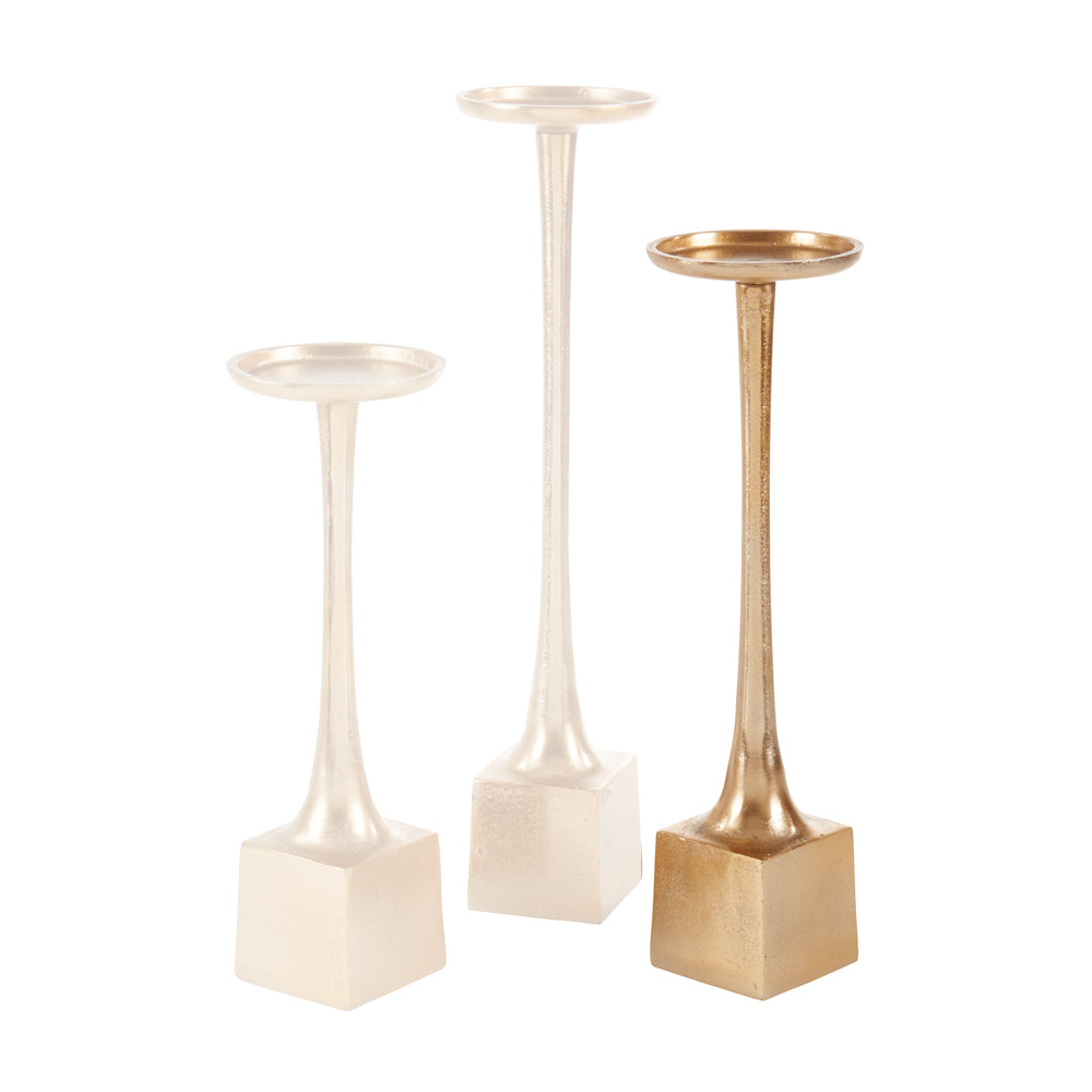 Three decorative candlestick holders with square bases and slender stems, shown on a plain white background. Two are warm off-white/ivory and one is metallic gold.