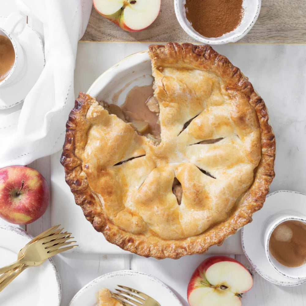 Vietri italian bakers pie dish with apple pie being cooked with it and a slice taken out, surrounded by apples and coffee cups on a marble surface.
