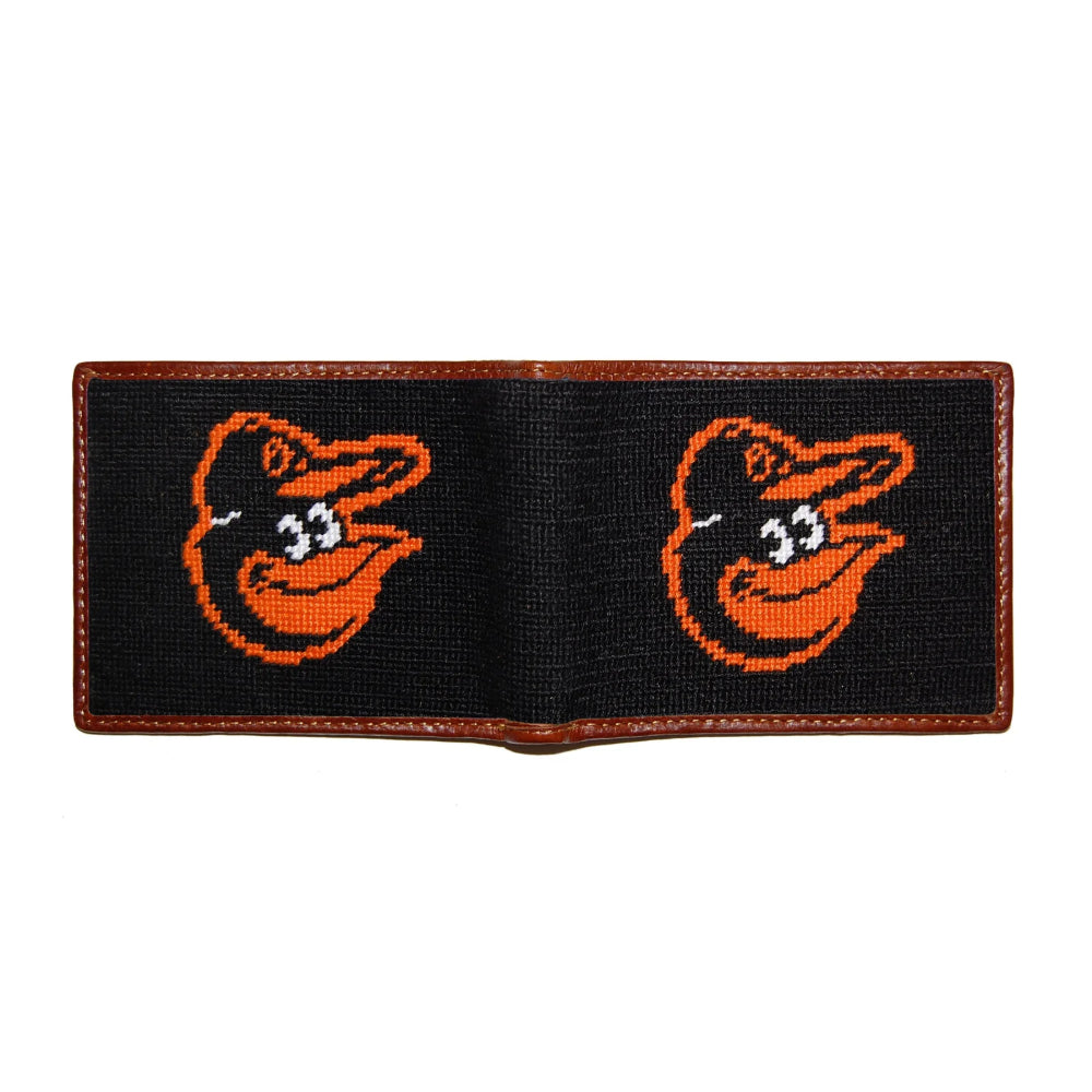 A black woven bifold wallet with brown leather trim, featuring two orange Baltimore Orioles bird logos embroidered on the front panels.