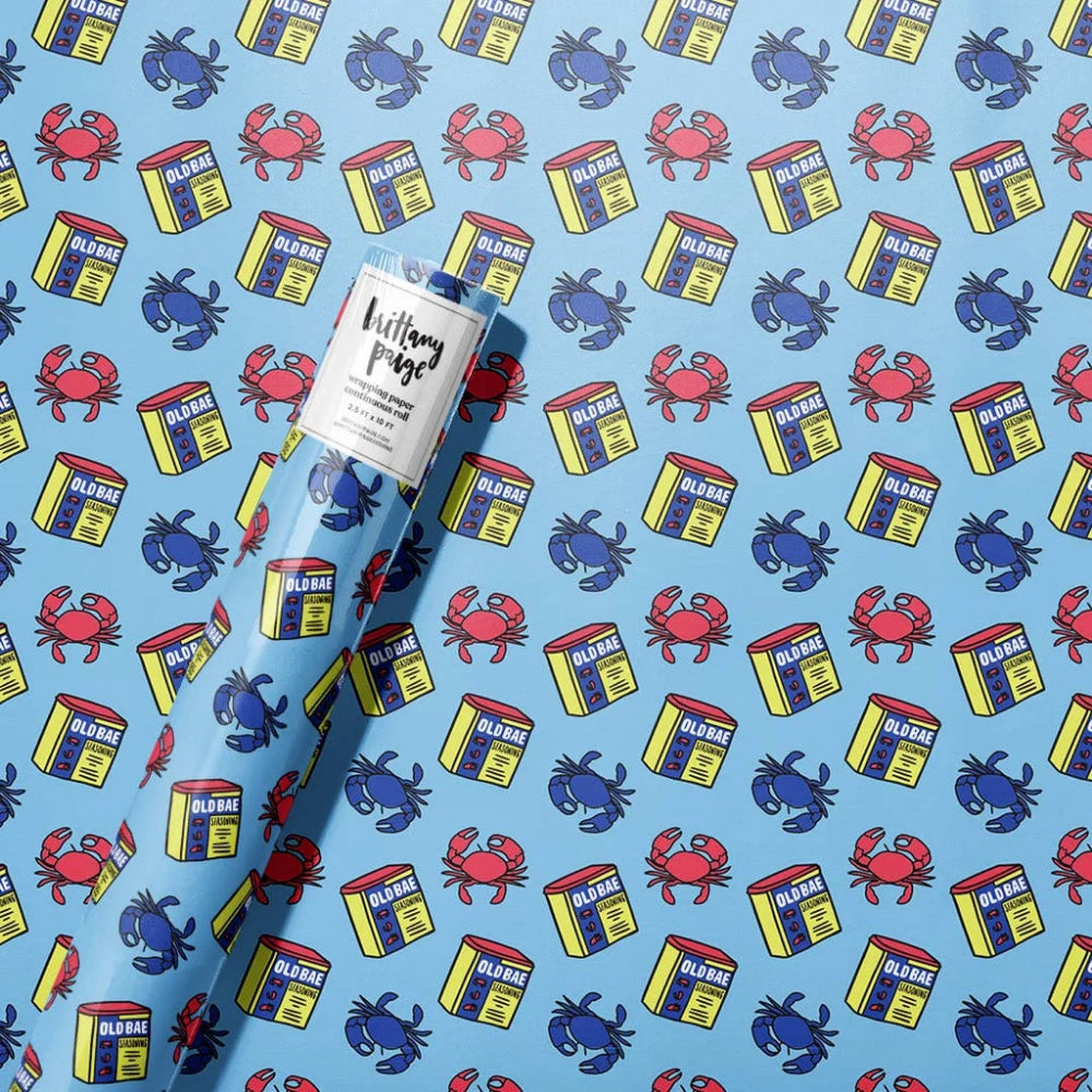 A roll of wrapping paper featuring a repeating pattern of blue and red crabs and yellow-and-blue Ollie Bag boxes on a light blue background, with a white paper label wrapped around the center.