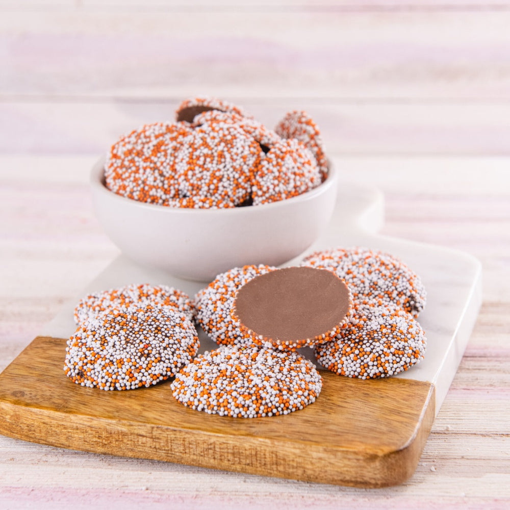 Nonpareils chocolates with orange and white sprinkles on a wooden board and white plate.