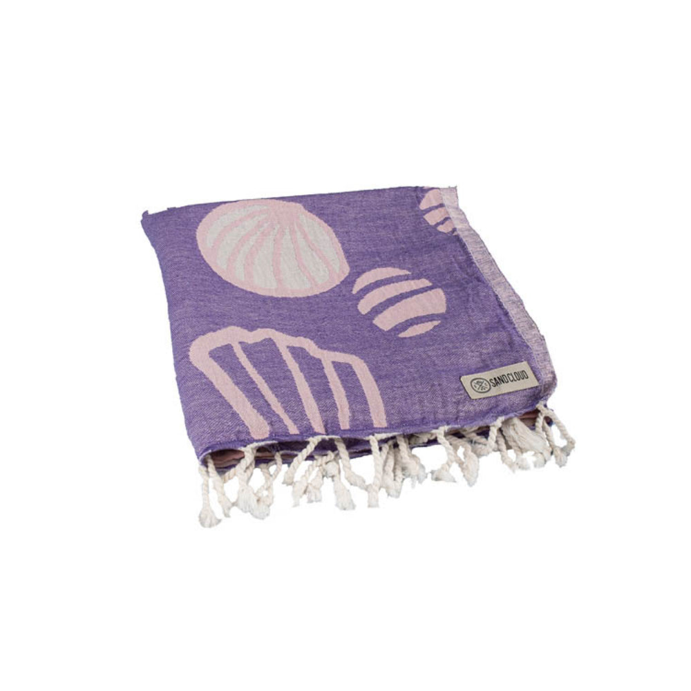 Folded purple and pink beach towel with a wave, shell,  and sea turtle pattern with a white tassel edge on a white background.