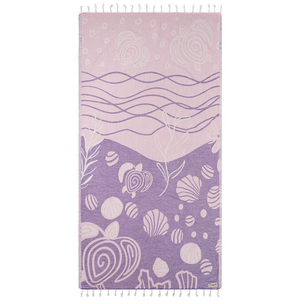 Side one of a purple and pink beach towel with a wave, shell,  and sea turtle pattern with a white tassel edge on a white background.
