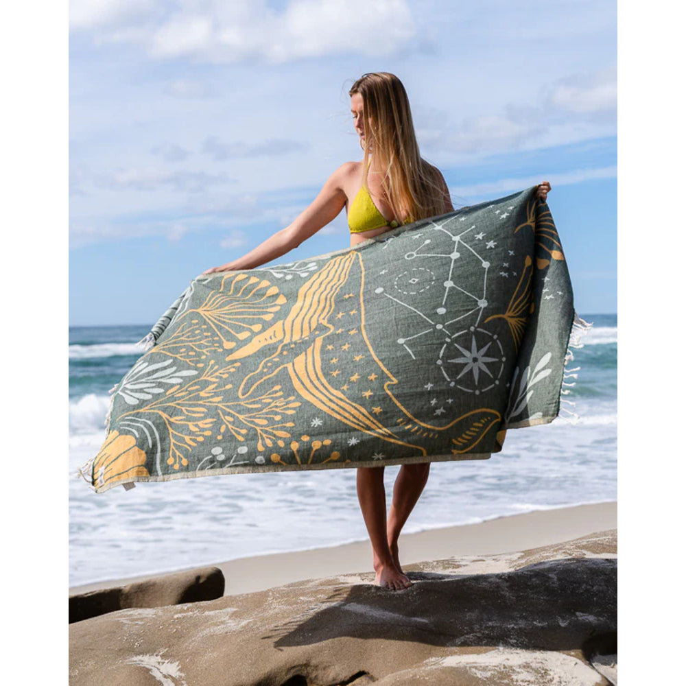 Woman holding a large blue and gold towel with celestial design on a beach.