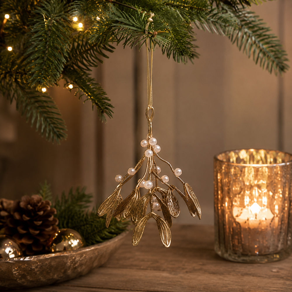 A gold mistletoe-shaped hanging ornament with small pearl-like berries is suspended from a Christmas tree branch. The scene is styled on a wooden surface with a lit candle in a glass holder, pinecones, and greenery in the background.