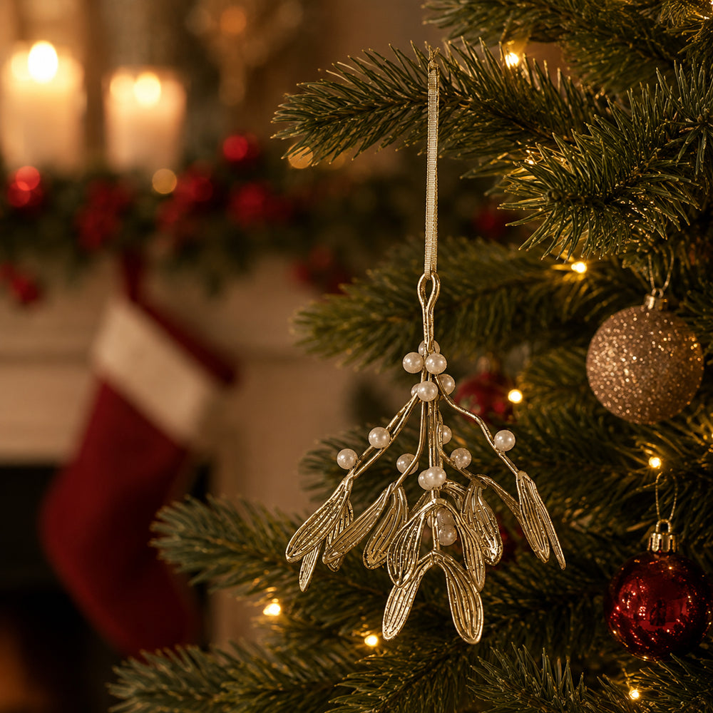 A silver mistletoe Christmas ornament with small white pearl-like beads hangs on a decorated Christmas tree branch, with warm holiday lights and a blurred red stocking in the background.