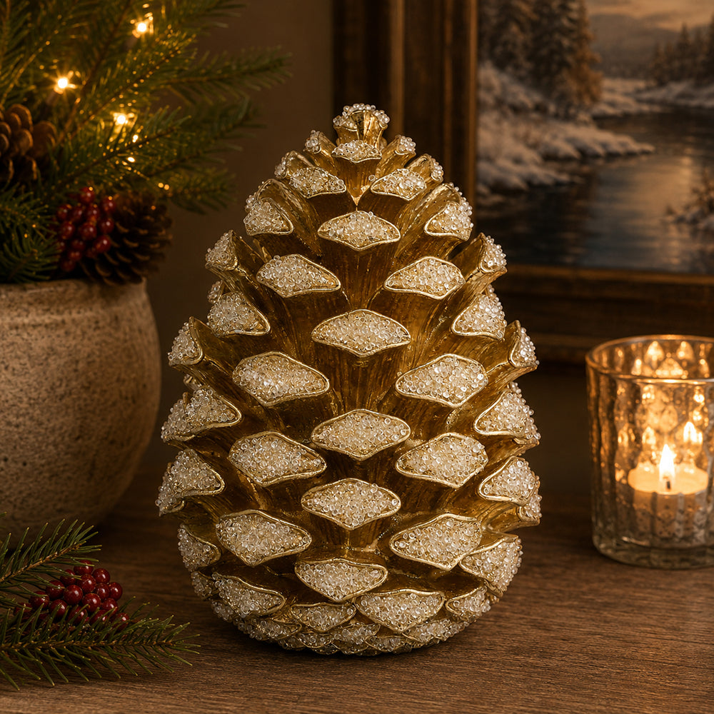 A decorative artificial pinecone ornament with textured gold and white glitter scales, displayed on a wooden surface in a cozy holiday setting with a lit candle and evergreen branches.
