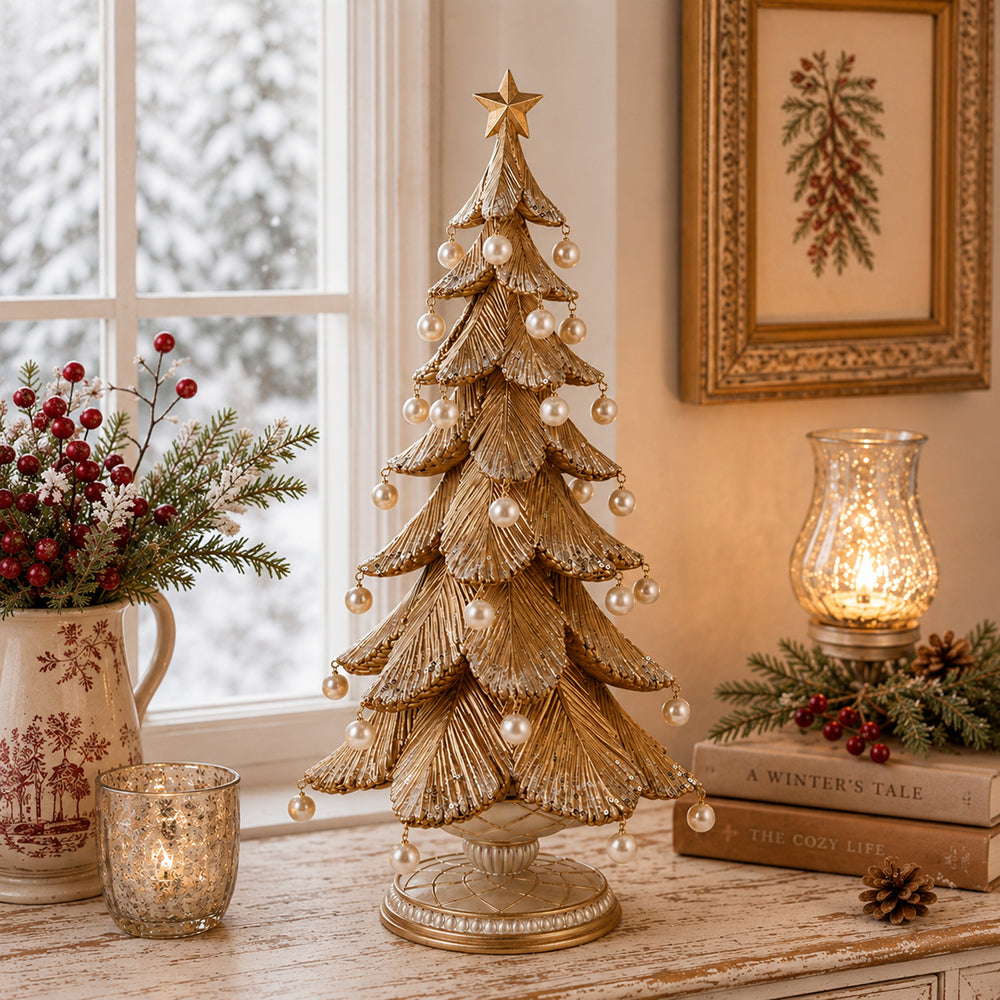 A decorative gold Christmas tree with a star topper and pearl-like ornaments stands on a distressed wooden table in a cozy room setting. The scene includes a lit candle holder, books, a floral pitcher, and a window with snowy outdoor scenery in the background.
