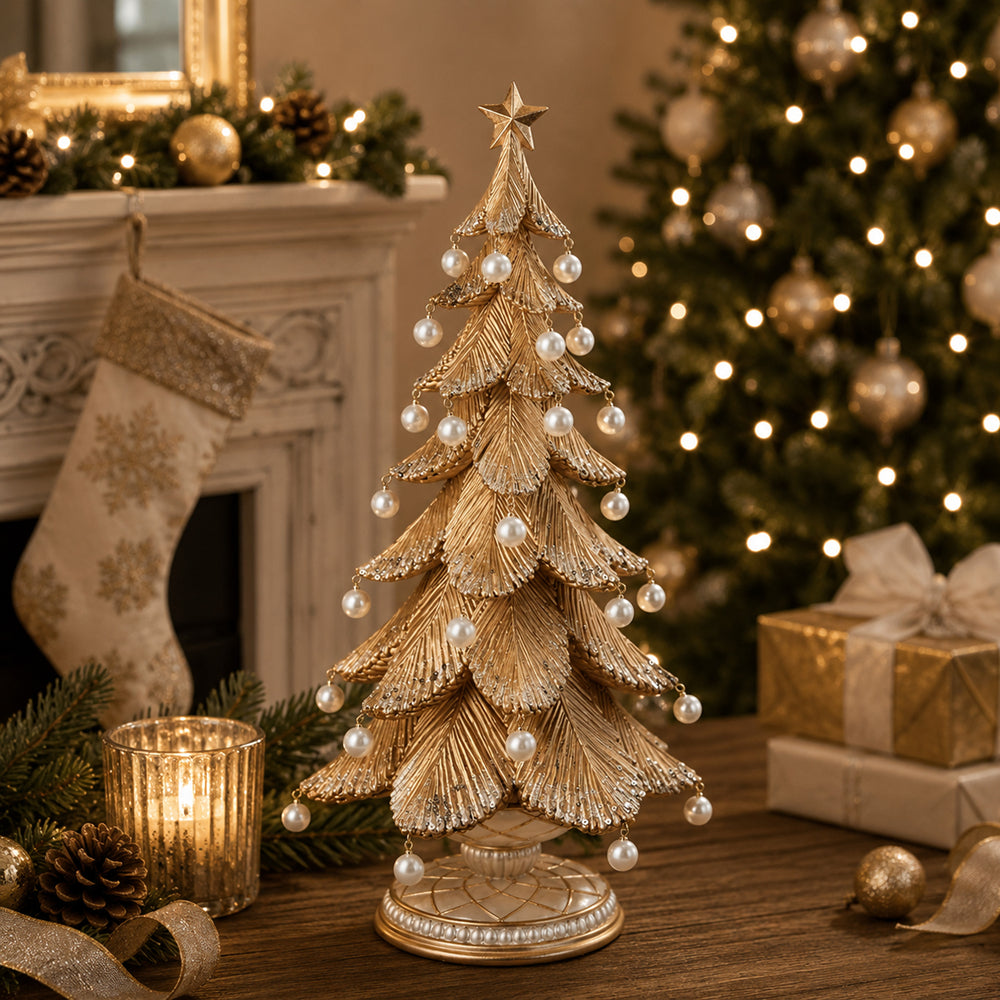 A decorative gold Christmas tree centerpiece with layered metallic leaves and hanging pearl-like ornaments, styled on a wooden table in a warm holiday setting with a lit fireplace, stockings, wrapped gifts, and a glowing Christmas tree in the background.