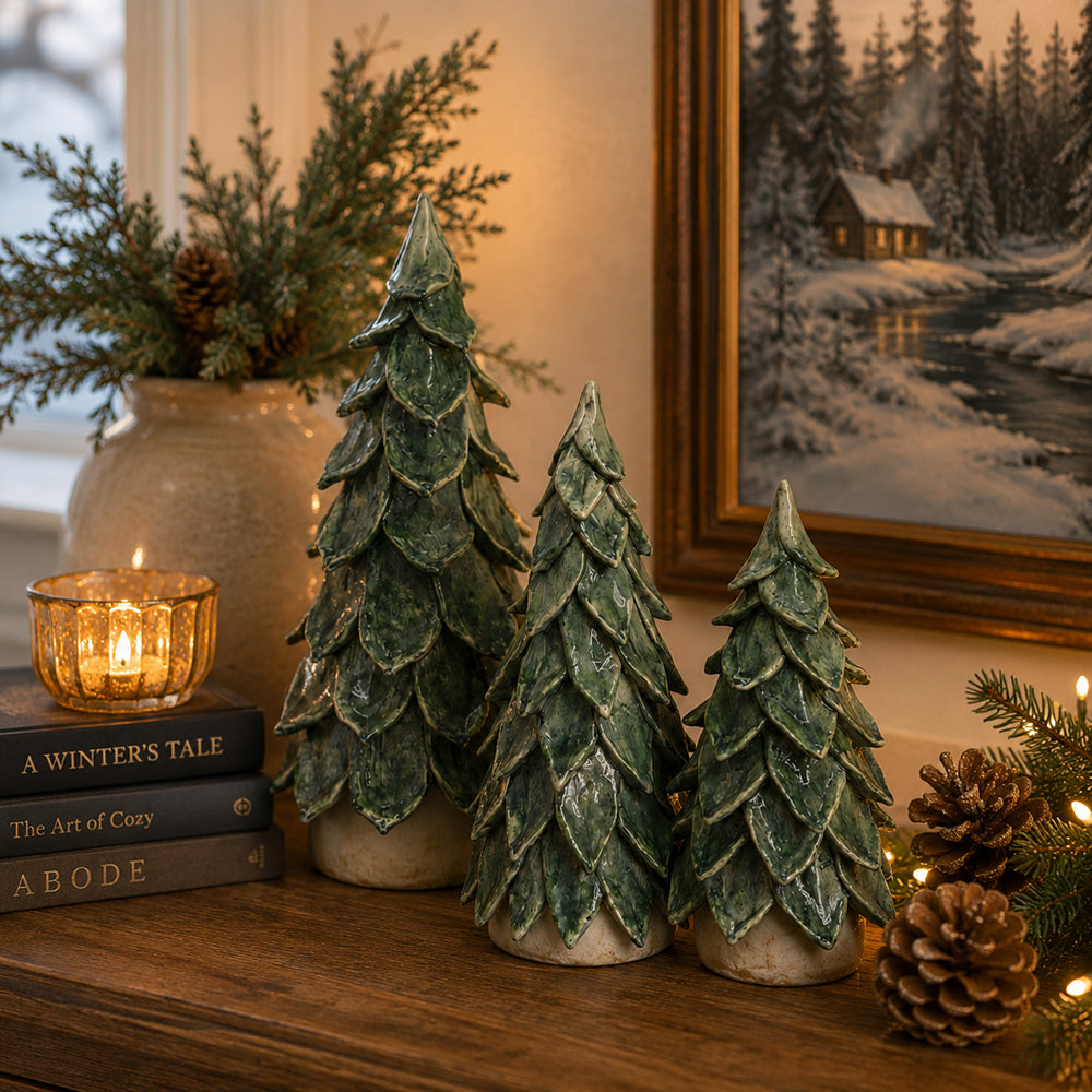 Three green pine tree ceramic ornaments with a textured, layered scale design, displayed on a wooden mantelpiece alongside a lit candle, books, pinecones, and a framed winter landscape painting.