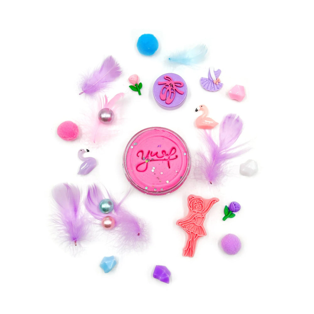 A flat-lay arrangement of colorful ballet-themed accessories and small toys on a white background, including a pink round tin with the word 'ballet' in script, purple and pink pom-poms, feathers, beads, a small flamingo figure, a ballerina silhouette, and other pastel decorative pieces.