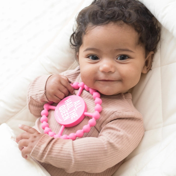 Baby holding a pink teething ring with text, lying on a white surface