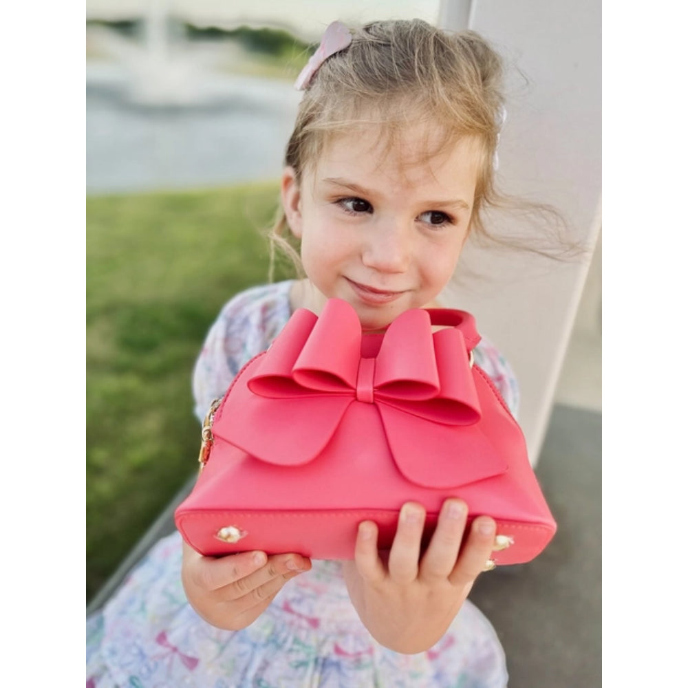 Young girl holding a pink handbag with a large bow outdoors.