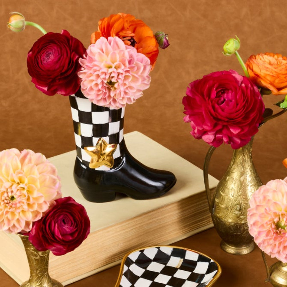 A decorative black-and-white checkered boot-shaped vase filled with flowers, styled on a book with matching floral arrangements and gold vases against a brown backdrop.