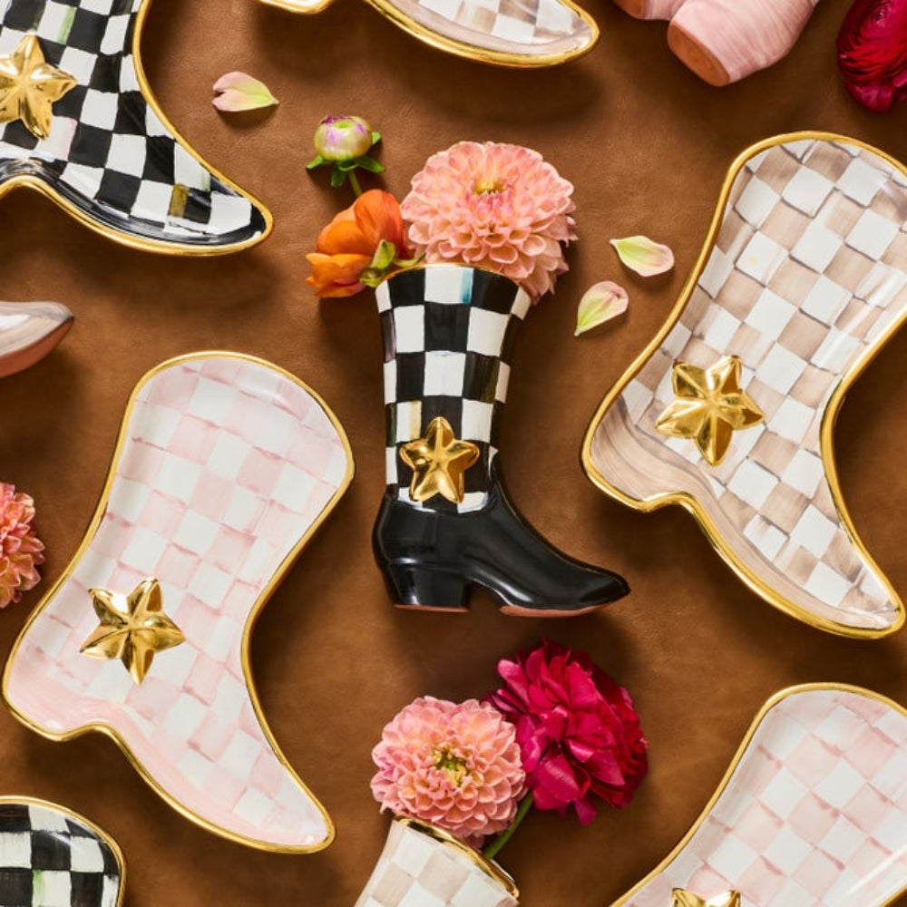 A styled flat-lay of ceramic boot-shaped plates on a brown surface, with one central plate shaped like a black-and-white checkered cowboy boot holding pink flowers, surrounded by other pastel and patterned plates.