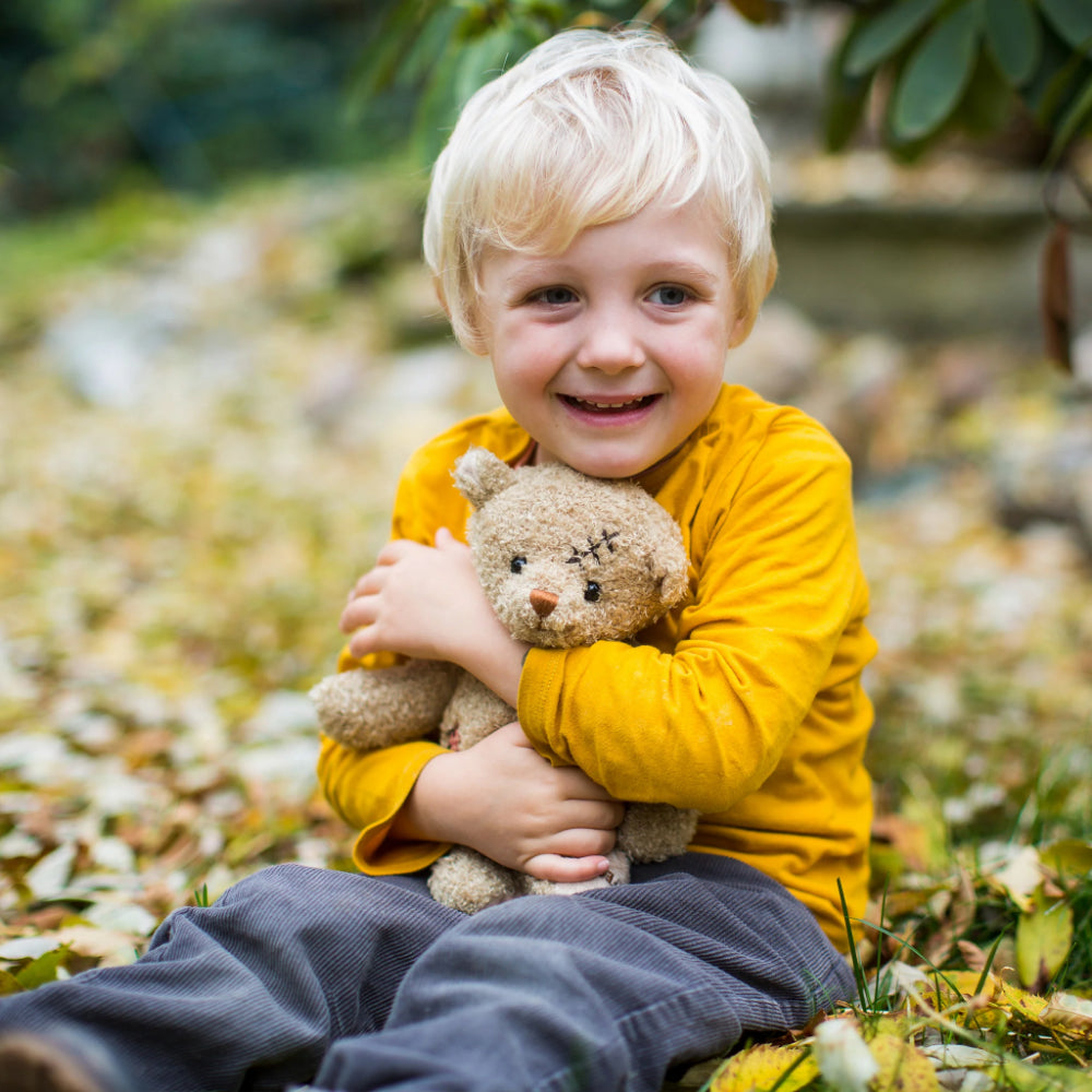 A beige stuffed bear with a scar on its head and a wound on its side, wearing a red and white checkered shirt, sitting upright with the phrase 'Nobody is Perfect' printed on its foot. Being hugged by a boy in a yellow shirt outside. 