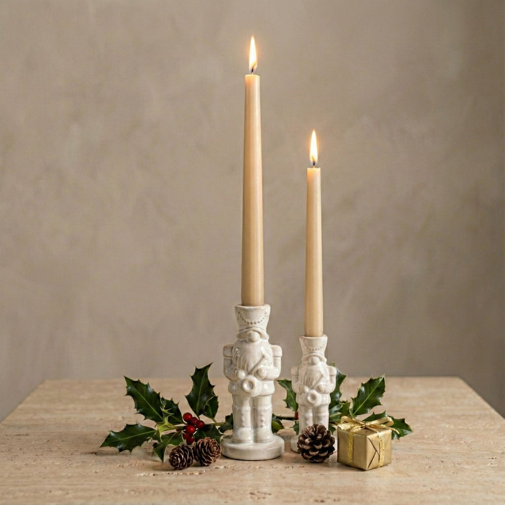 Two lit cream taper candles in white nutcracker-shaped holders, styled on a rustic wooden surface with holly, pinecones, and a small gold gift box against a neutral beige background.
