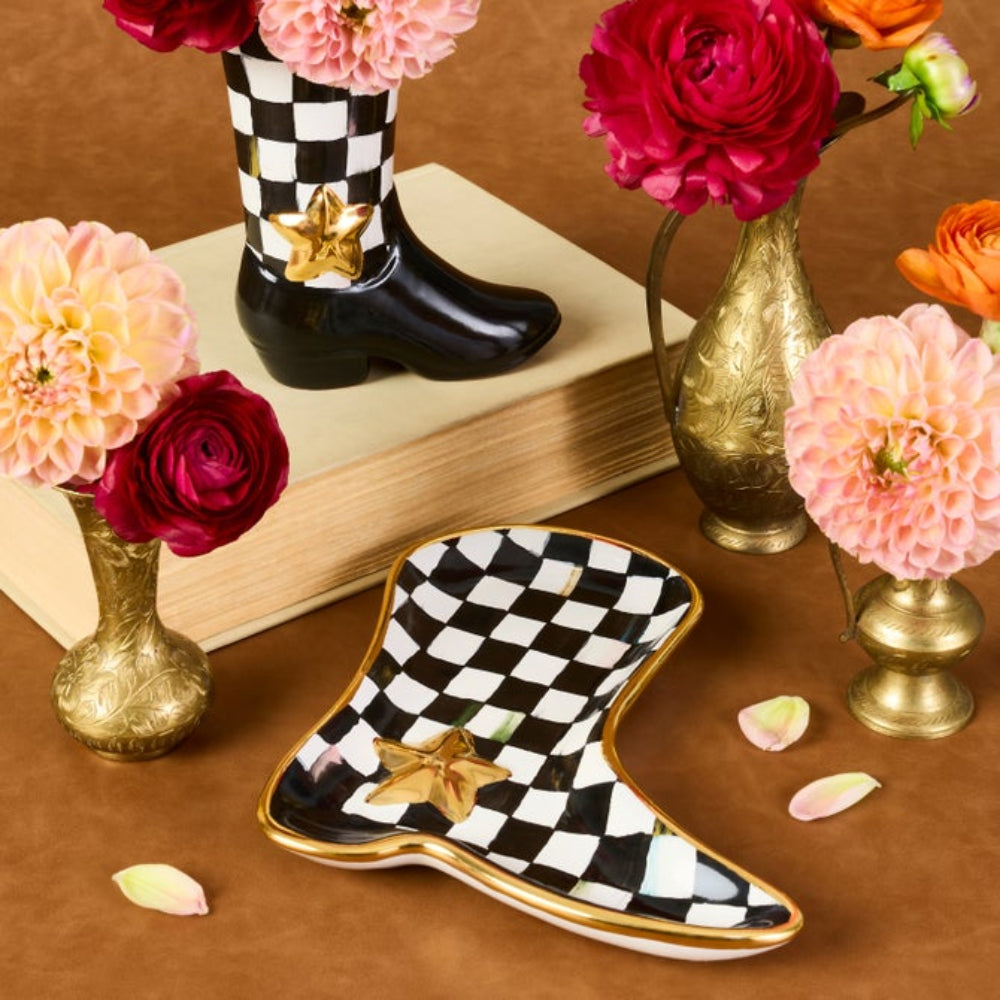 A decorative boot-shaped dish with a black and white checkerboard pattern and a gold star detail, styled on a brown surface with flowers and gold vases.