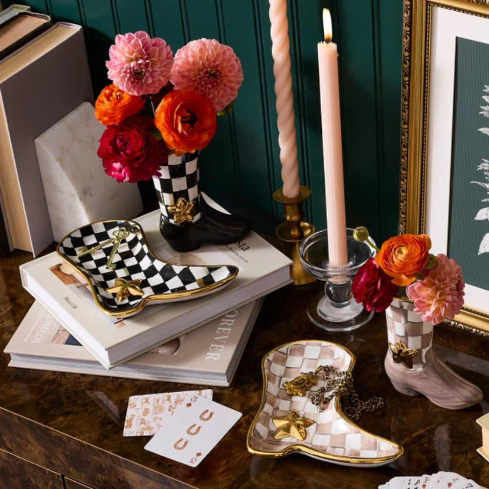 A styled tabletop scene with two cowboy boot-shaped ceramic dishes, one black-and-white checkered and one beige-and-white checkered with gold trim, holding flowers and candles on a dark wood surface against a deep green paneled wall.