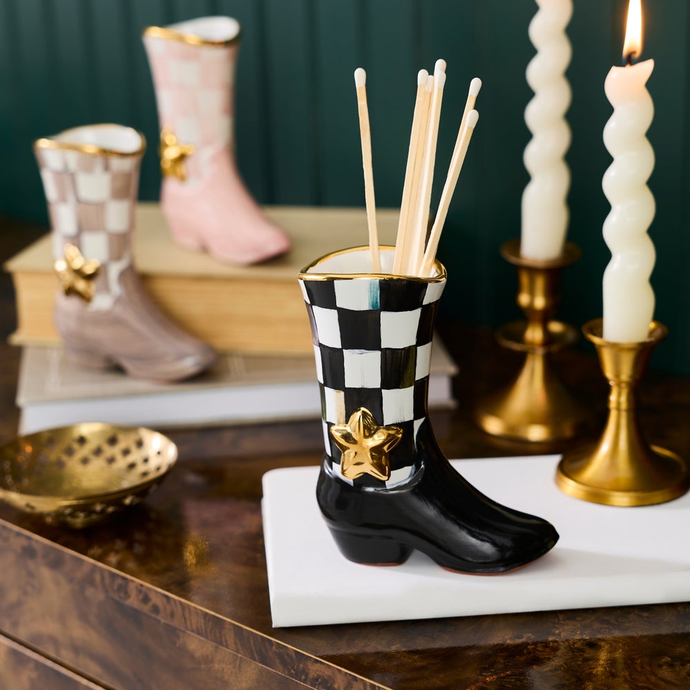 A ceramic incense holder shaped like a cowboy boot, featuring a black and white checkerboard pattern on the shaft, a glossy black toe, and a gold star accent, shown holding incense sticks on a tabletop.