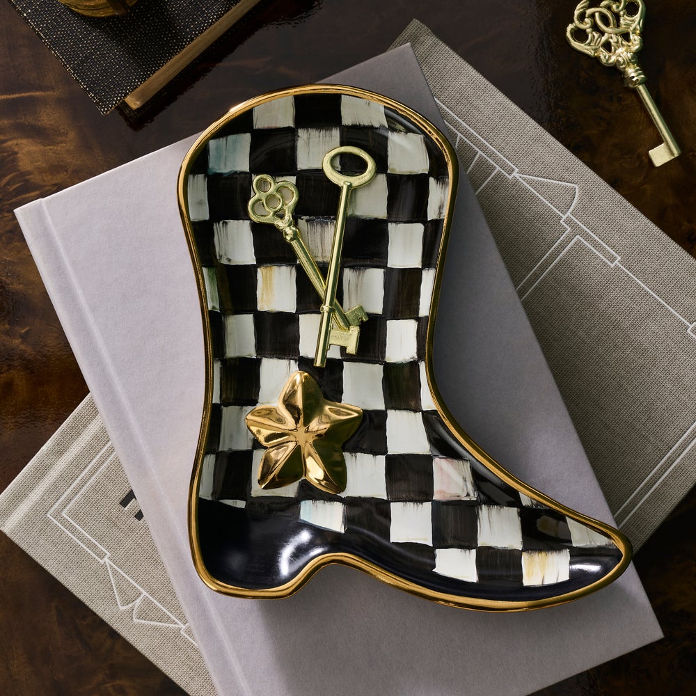 A decorative boot-shaped tray with a black and white checkerboard pattern and gold trim, styled on top of books with two ornate gold keys resting inside it.