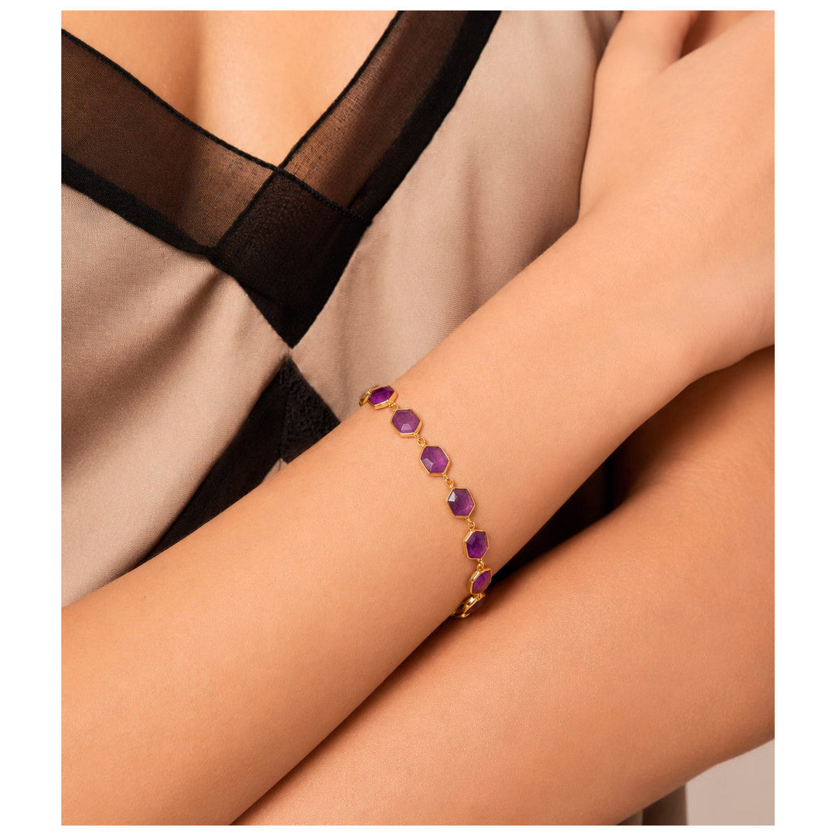 A gold bracelet with a row of purple oval gemstones worn on a model's wrist, shown against a beige and black top.