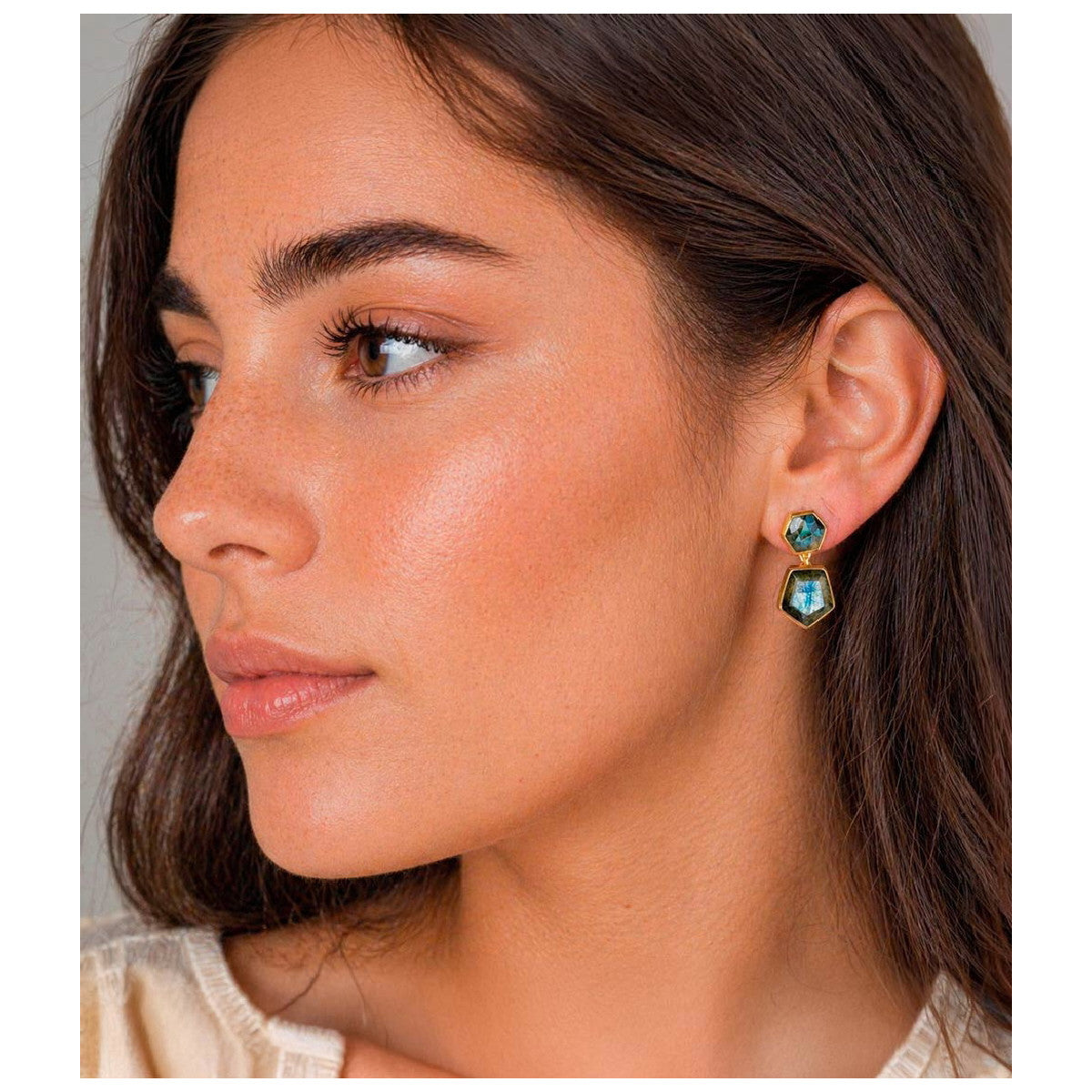 A close-up of a woman wearing a gold drop earring with two stacked oval green gemstones, shown in profile against a neutral background.