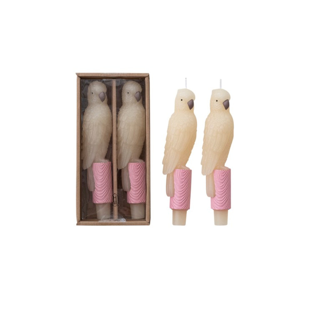 Two parrot-shaped candles with pink bases in a cardboard box on a white background