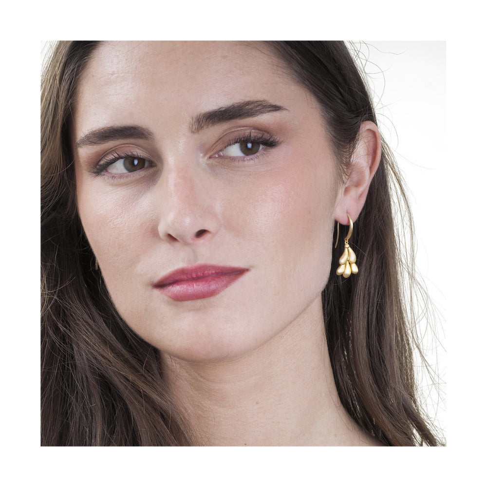 A close-up portrait of a woman wearing a small gold dangle earring with three clustered leaf-like drops, set against a plain white background.
