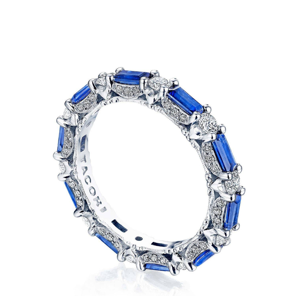 A silver band ring featuring baguette-cut royal blue sapphires and small clear stones, shown on a clean white background. The inner band is engraved with the brand name 'TACO'.
