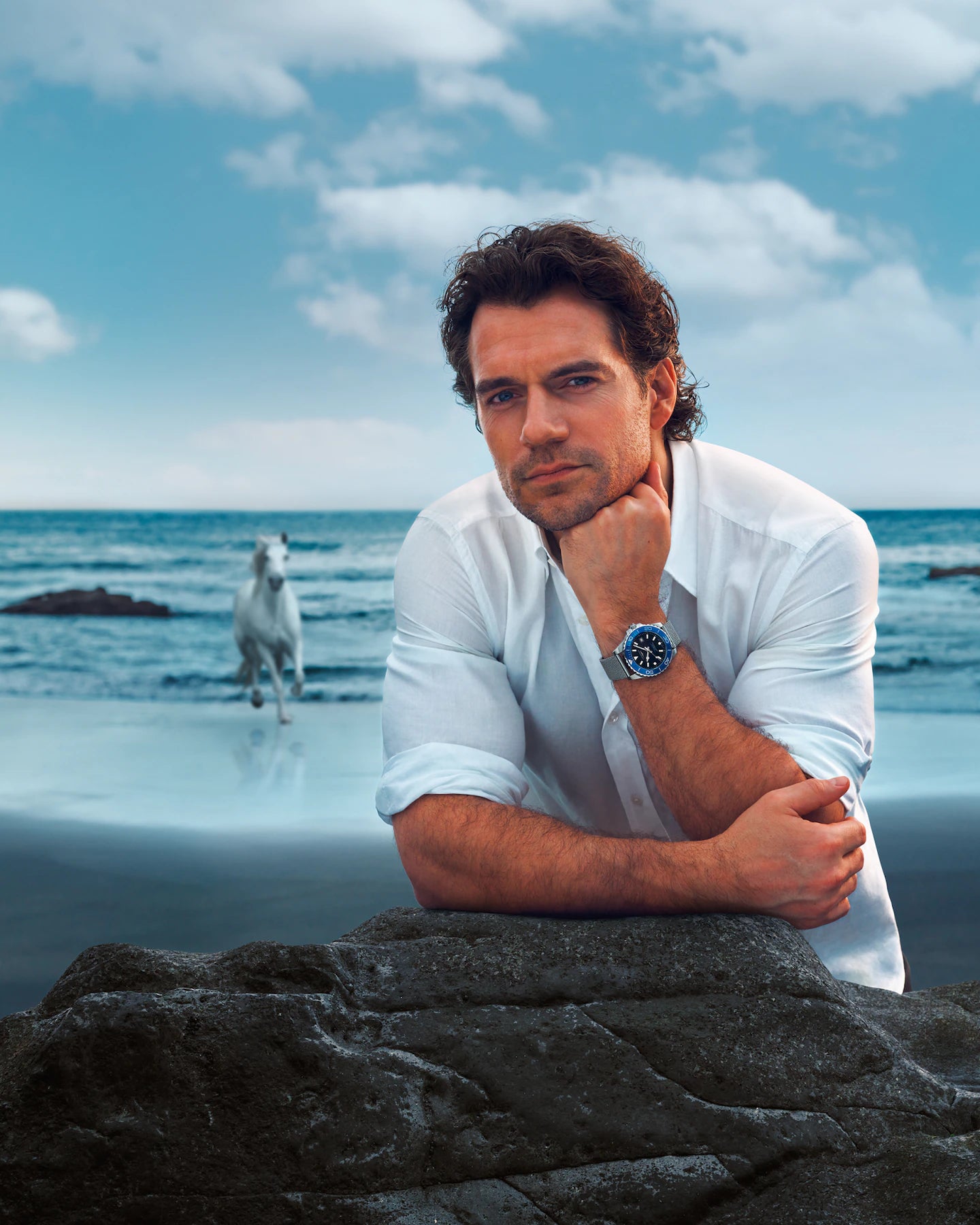 Man in a white shirt sitting on a rock by the ocean with a dog running in the background