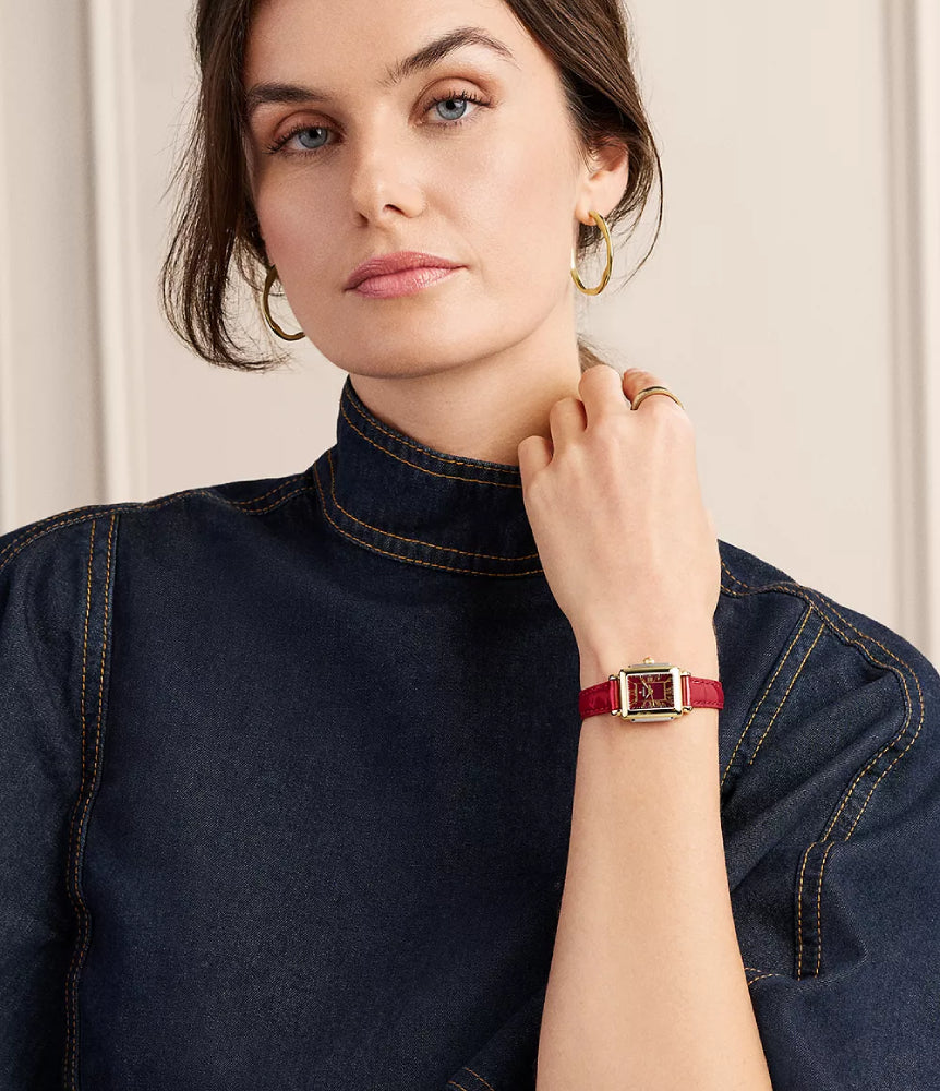 A woman wearing a red leather strap watch with a gold-tone rectangular case, styled with a dark denim mock-neck top and gold hoop earrings.
