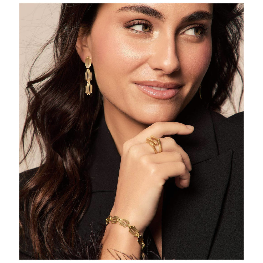 A close-up lifestyle shot of a woman wearing gold jewelry, featuring a gold link earring, a gold ring, and a gold bracelet, paired with a black blazer against a neutral beige background.