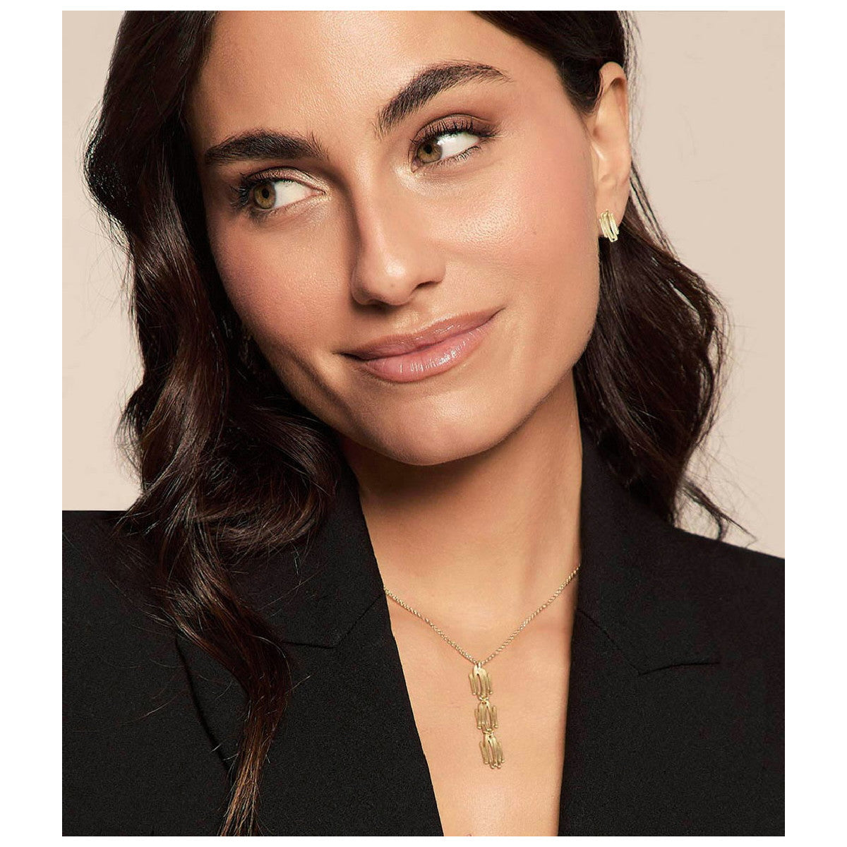 A woman with dark wavy hair wearing a delicate gold pendant necklace and a small gold stud earring, dressed in a black blazer against a solid beige background.