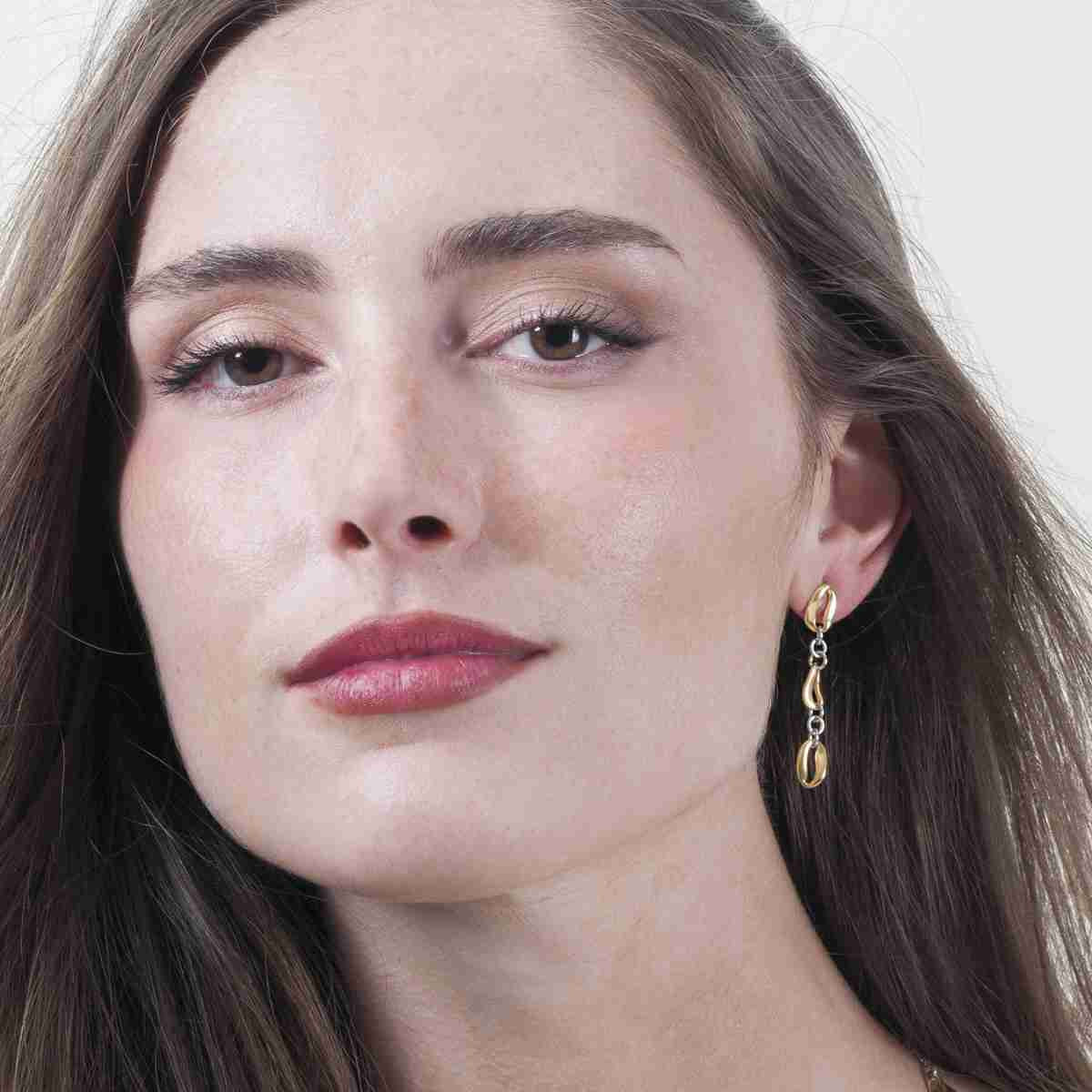 A close-up portrait of a woman wearing a delicate gold dangle earring with a small chain link and teardrop-shaped drop, against a clean light background.
