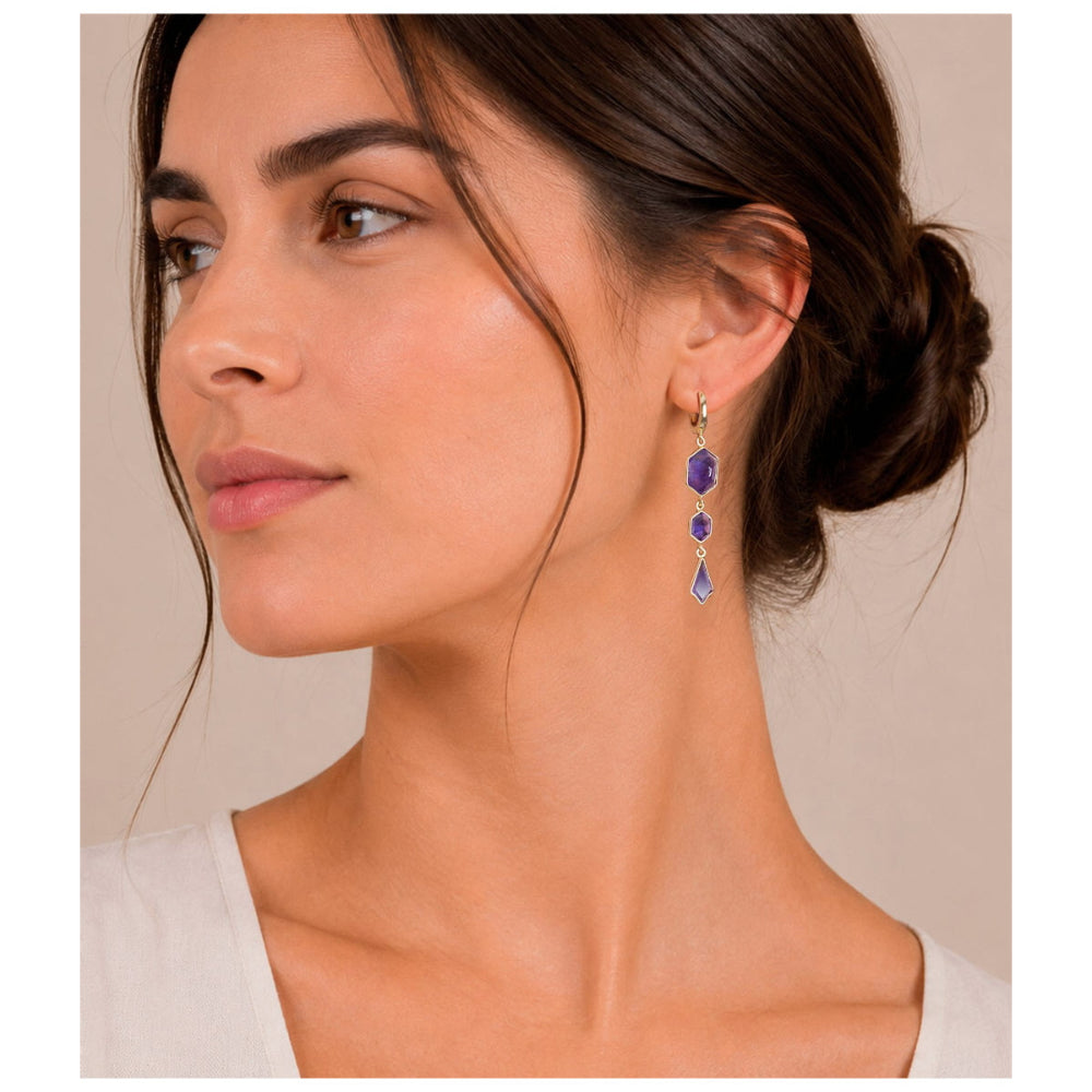 A woman in profile wearing a delicate gold dangle earring with three purple gemstone drops against a soft beige background.