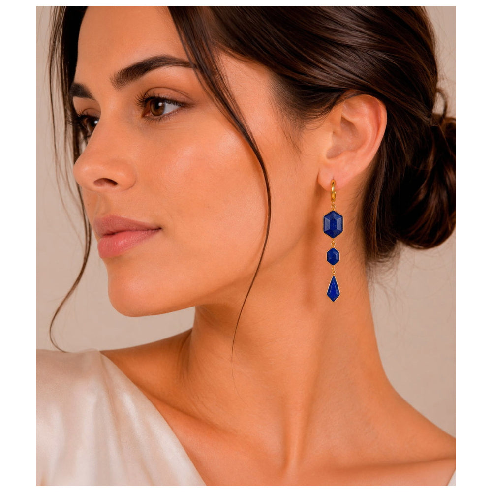 A close-up of a woman wearing a long drop earring with three faceted blue gemstones and gold-toned metal findings.