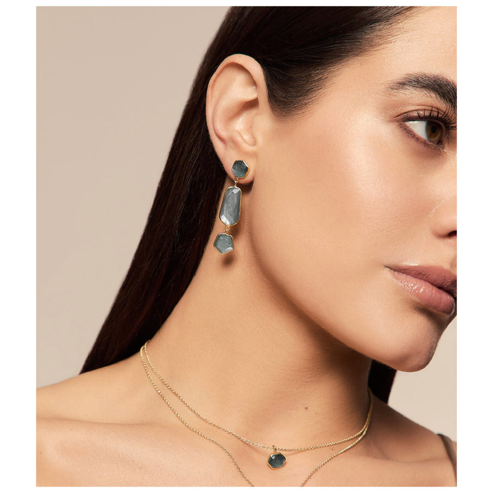 A close-up of a woman wearing a matching jewelry set, featuring a gold drop earring with three stacked rectangular gray-blue stones and a delicate double-strand gold necklace with a small oval gray-blue stone pendant, set against a solid beige background.