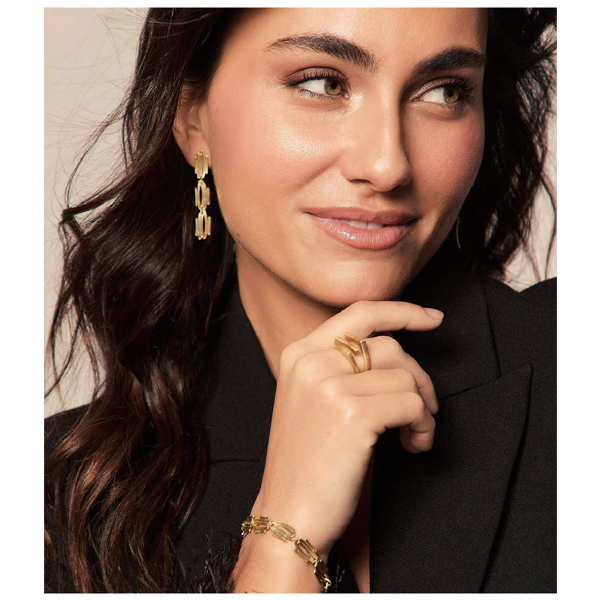A close-up lifestyle shot of a woman wearing a gold geometric link drop earring, a matching gold ring, and a gold link bracelet, paired with a black blazer.